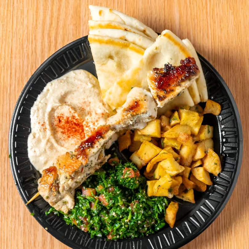 Taza Grill's chicken shish tawook with hummus, pita, tabbouleh, and seasoned potatoes on a black plate.