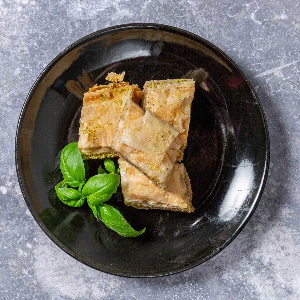 Taza Grill's baklava dessert, golden brown with pistachio crumbles and fresh basil on a black plate.
